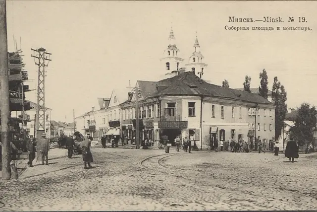 Old postcard of a cobbled square with tram tracks, a corner house with shops and the twin towers of the Holy Spirit church behind