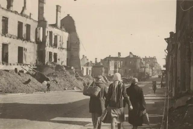 Three townspeople with baskets walking down a street lined with burnt-out, roofless buildings and heaps of rubble