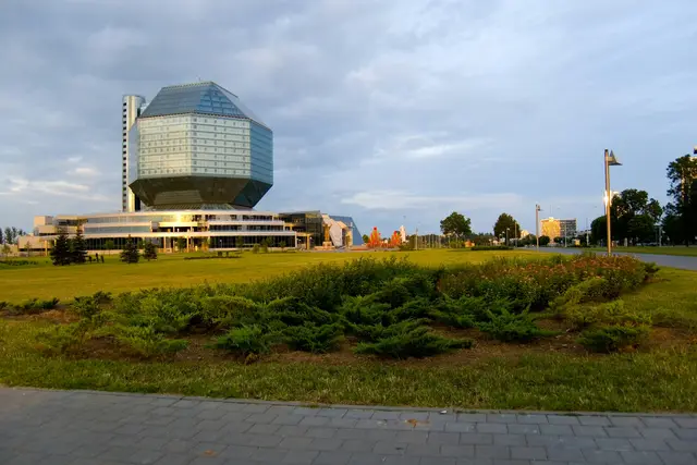 The diamond-shaped glass National Library of Belarus in evening sun beyond a lawn and juniper beds